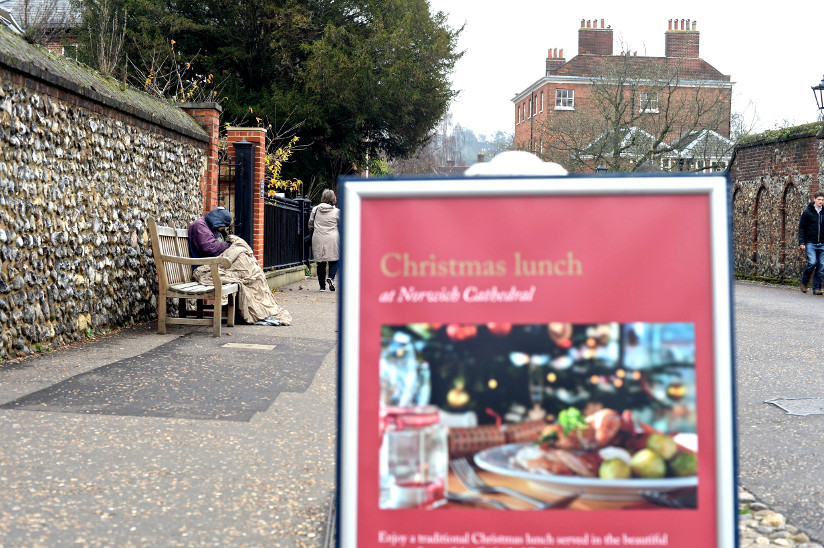 Photo of homeless man sitting on a bench behind an advertisment for Christmas lunch